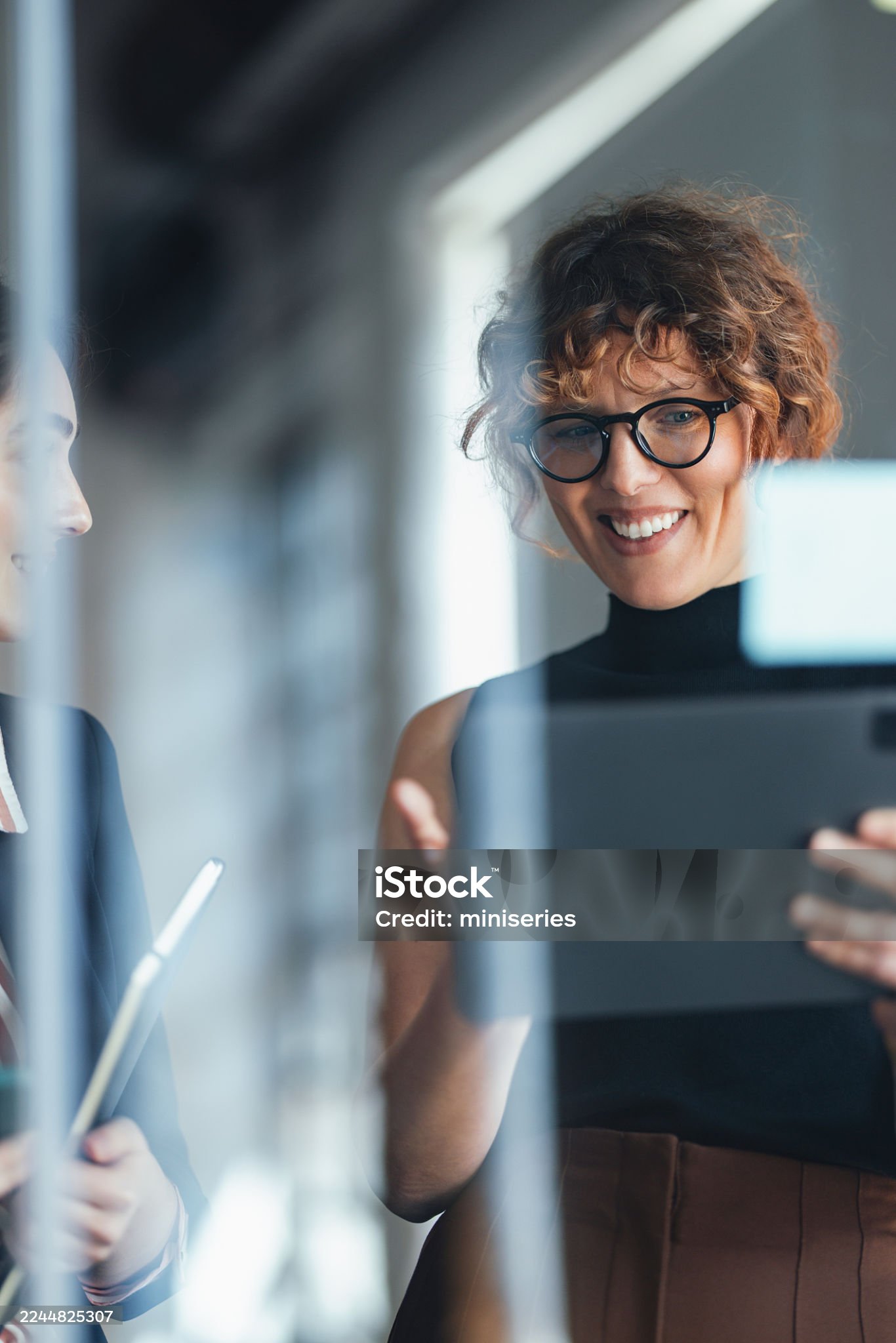 A confident woman in a black top and glasses smiles while holding a tablet in a modern coworking office, suggesting collaboration, productivity, and professional energy in a dynamic work environment.